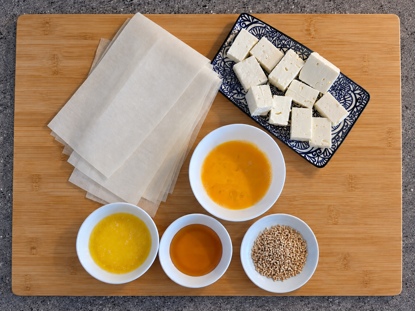 Filo pastry, feta pieces, beaten egg, melted butter, honey and sesame seeds arranged on a wooden chopping board