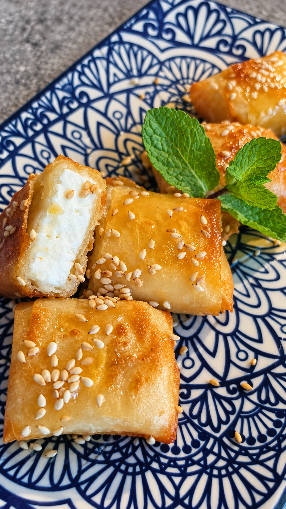 Crispy filo-wrapped feta bites with honey and sesame on a patterned plate, with one cut open to reveal the feta centre